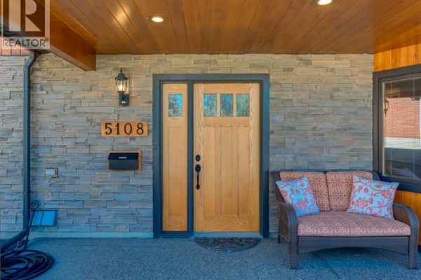 Close-up of a wooden front door with glass panels, stone siding, and a small outdoor seating area. - 5108 Brockington Road Nw, Calgary