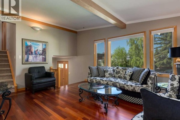 Living room with sofas, a black armchair, hardwood floors, a staircase, and a view of trees through the windows. - 5108 Brockington Road Nw, Calgary