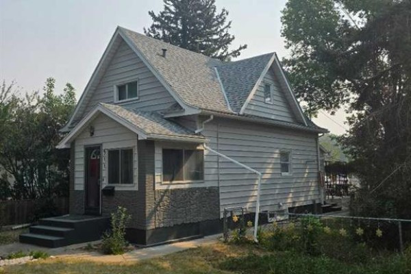 Side view of a light blue and grey two-story house with a peaked roof and green trees. - 511 12c Street N, Lethbridge