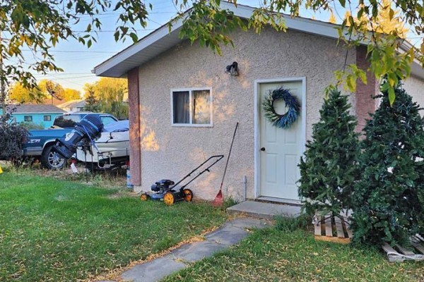 Exterior of a small tan stucco building with a white door, window, lawnmower, and two artificial Christmas trees. - 5111 53 Street, Mannville