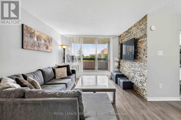 Living room with large grey sectional sofa, coffee table, and balcony access with vertical blinds. - 512 - 1615 Bloor Street, Mississauga
