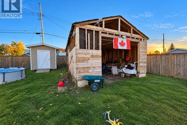 Unfinished wooden garage with tools, a Canadian flag, and a wheelbarrow in a grassy backyard. - 5122 45 Avenue, Spirit River