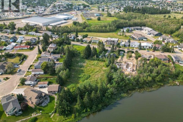 Aerial shot showcasing the lakefront property with surrounding houses, undeveloped land, and distant commercial buildings. - 5128 & 5124 56 Street, Innisfail