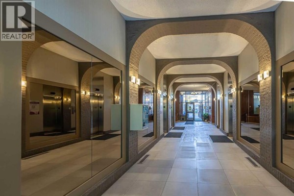 A long apartment building hallway with arched doorways, mirrored walls, and elevator entrances. - 513, 4944 Dalton Drive Nw, Calgary