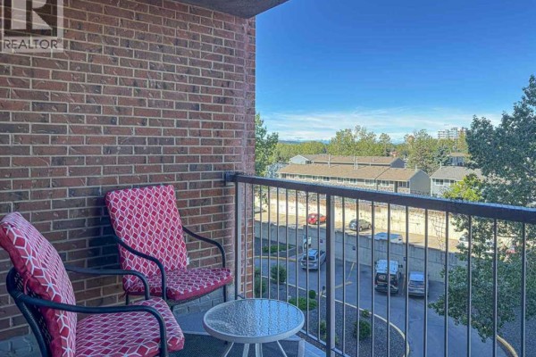 A small balcony with two chairs, a table, and a view of a parking lot and trees. - 513, 4944 Dalton Drive Nw, Calgary