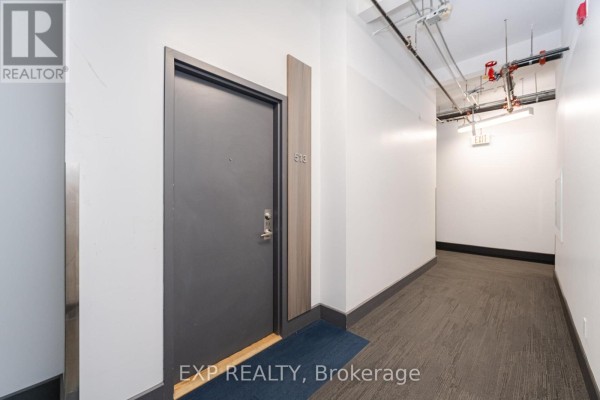 Hallway leading to apartment 513 with a grey door and industrial pipes overhead. - 513 - 955 Queen Street W, Toronto