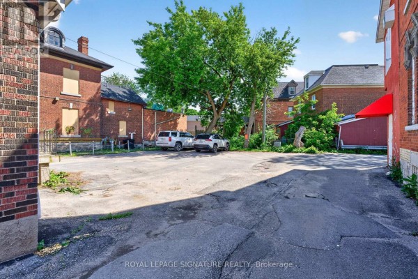 A paved lot behind brick buildings with parked cars and green trees overhead. - 514 Princess Street, Kingston