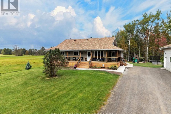 Front exterior of a beige house with a large wooden wrap-around porch and gray roof. - 5140 Arena Boundary Road, Frontenac