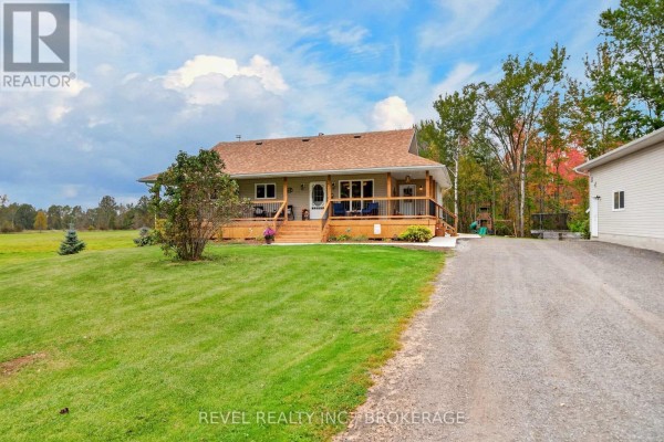 Long shot of the front of a beige home with a covered porch and surrounding green lawn. - 5140 Arena Boundary Road, Frontenac