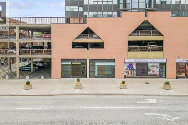 Exterior view of a commercial building showing storefronts, parking garage, and surrounding street. - 515 Riverside Drive West Unit# 104 A, Windsor