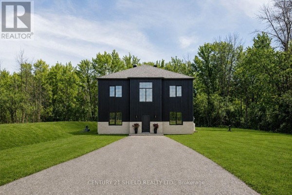 View of the modern house from the end of a long gravel driveway, with green grass and trees. - 5153 Archer Road, South Dundas