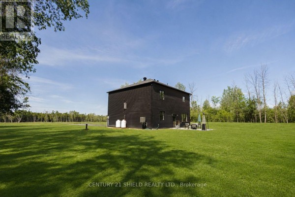 Side view of the dark modern home surrounded by a large grassy field and trees. - 5153 Archer Road, South Dundas