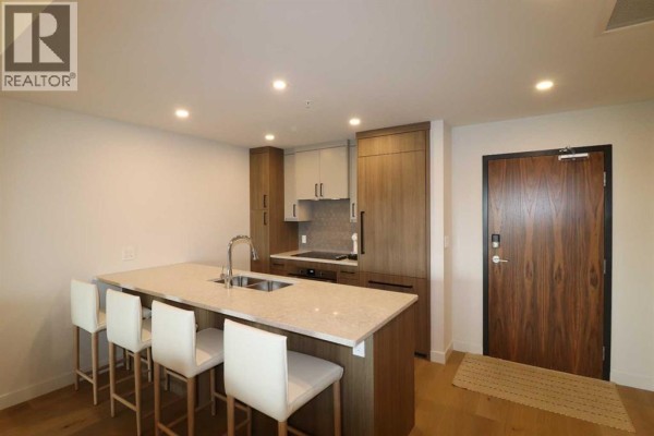 Kitchen island with bar stools, facing a built-in stovetop, oven, and wood grain cabinetry. - 518, 102 Scenic Drive N, Lethbridge