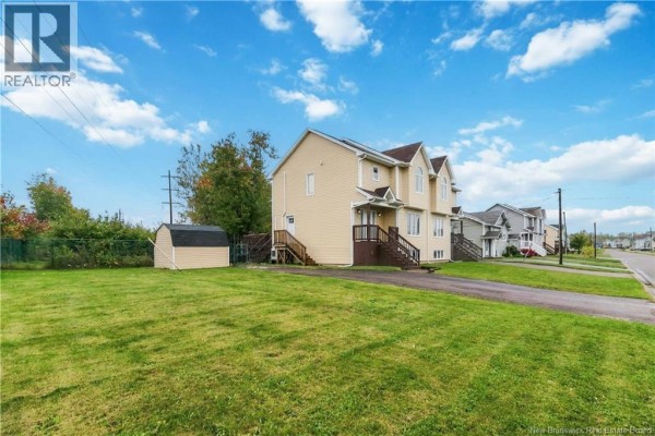 Wide exterior view of a beige townhouse with a green lawn, driveway, and neighboring townhouses. - 52 Coriander Street, Moncton