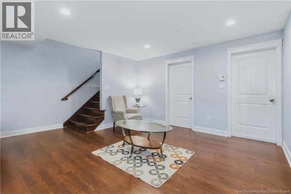 Living room with light blue walls, wood floors, a beige armchair, glass coffee table, and patterned rug. - 52 Coriander Street, Moncton