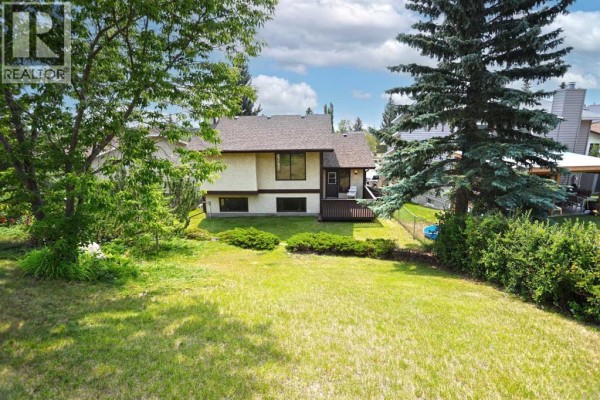 Backyard view of a house with a deck, grassy lawn, and mature trees. - 52 Edgeland Road Nw, Calgary