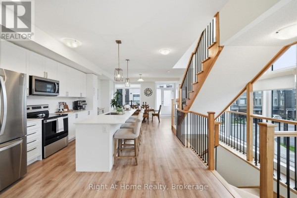 Modern kitchen with white cabinetry, island with bar stools, and adjacent dining area with stairs. - 52 Emmas Way, Whitby