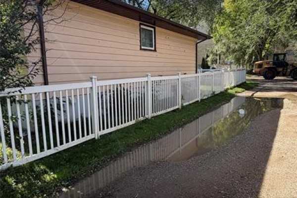 White vinyl fence along a gravel driveway with a large puddle and construction equipment in the background. - 521 17th Street, Brandon