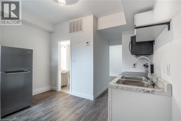 Kitchenette area with stainless steel refrigerator, double sink, stovetop, and doorway to bathroom. - 524 King Street E Unit# 3, Hamilton