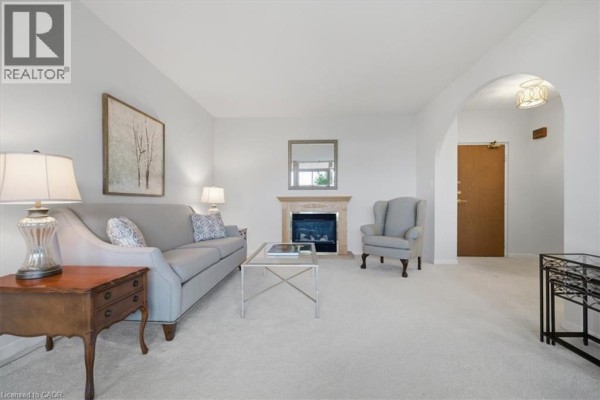 Living room with sofa, armchair, fireplace, and view of the hallway entrance and dining area. - 5250 Lakeshore Road Unit# 401, Burlington