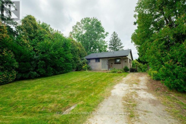 A single-story house with a light brick exterior, a gravel driveway, and a lawn surrounded by trees. - 526 Stafford Drive, Oakville