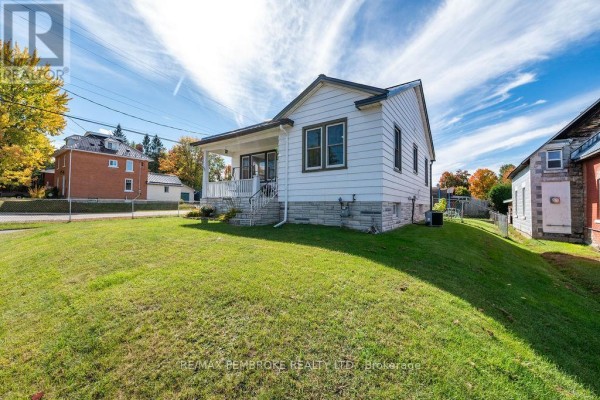 Side view of a white house with a metal roof, stone foundation, and green lawn on a sunny day. - 53 Doran Street, Petawawa