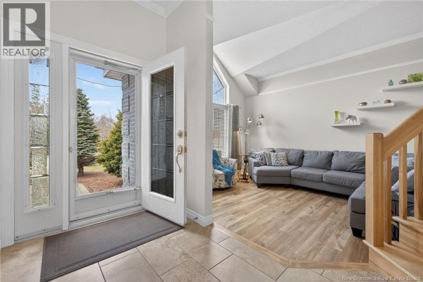 Interior view showing a living room with a grey sectional sofa, wooden staircase, and dark wood cabinets in the kitchen. - 53 Rue Mallet, Haut Shippagan