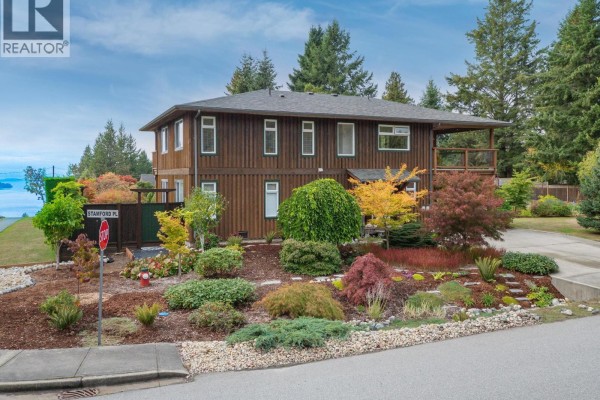 Wood-sided house with a well-landscaped front yard featuring colorful shrubs and trees, and a distant water view. - 5301 Stamford Place, Sechelt
