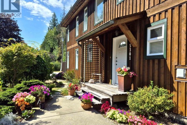 Front entrance of a wood-sided house with a covered porch, wooden steps, and potted flowers. - 5301 Stamford Place, Sechelt