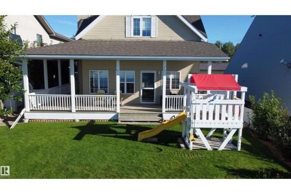 Rear exterior of a beige house with a covered porch, lawn, and children's play structure. - 5309 51 Av, Rural Lac Ste  Anne County