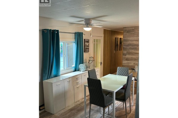 Dining area with a white buffet, glass-top table, chairs, and teal curtains. - 5317 Freda Avenue, Kamloops