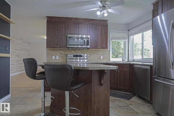 Kitchen island with a granite countertop and bar stools, next to stainless steel appliances. - 53212 Rge Road 14, Rural Parkland County
