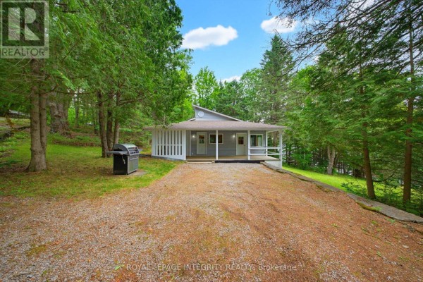 Gray cottage exterior with a barbecue on the gravel driveway, surrounded by trees. - 534 School House Lane, Greater Madawaska
