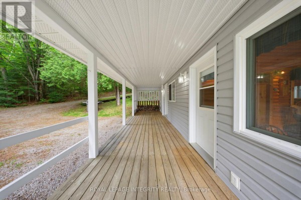 Covered wooden porch with white railings and gray siding, overlooking a wooded area. - 534 School House Lane, Greater Madawaska