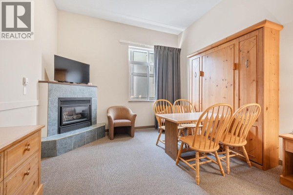 Studio apartment dining area with a light wood table and four chairs next to a fold-down bed and fireplace. - 5340 Big White Road Unit# 129, Big White
