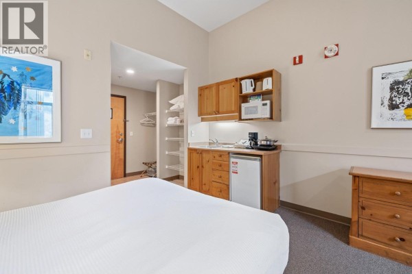 Studio apartment with a bed in the foreground, a compact kitchenette with wood cabinets, and a hallway view. - 5340 Big White Road Unit# 129, Big White