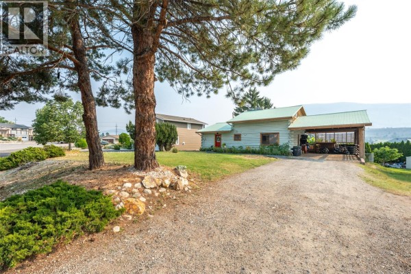 Gravel driveway leading to a light blue log home with a green metal roof and a covered carport. - 538 Lakeview Place, Oliver