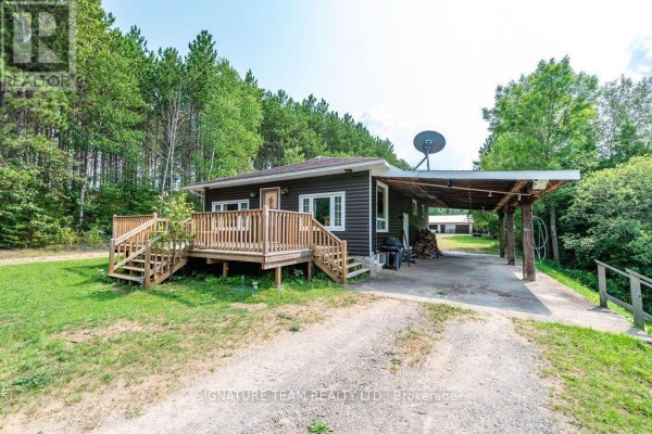 Side view of a dark brown bungalow with a wooden deck, carport, and BBQ, set against a backdrop of pine trees. - 538 Lenser Road, Bonnechere Valley
