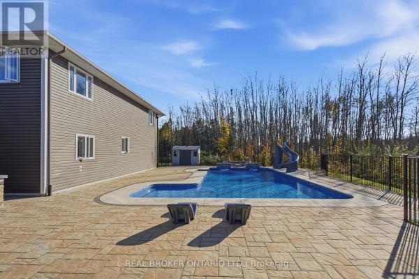 View of a swimming pool and patio with stone accents and a grey siding house with trees in the background. - 5381 Desmarais Road, Greater Sudbury