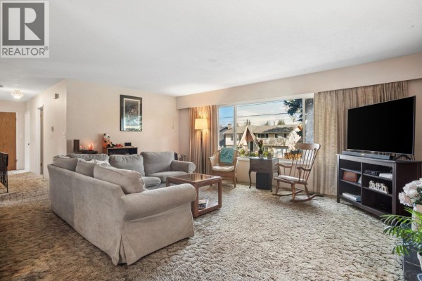 Living room with beige carpet, large window, and seating arrangement facing the television. - 5392 Maple Crescent, Delta