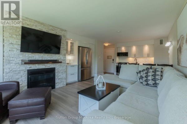 Living room with a fireplace, television, sofa, and a view of the kitchen. - 54 - 54-305 Deerhurst, Summit Lodge Drive, Huntsville