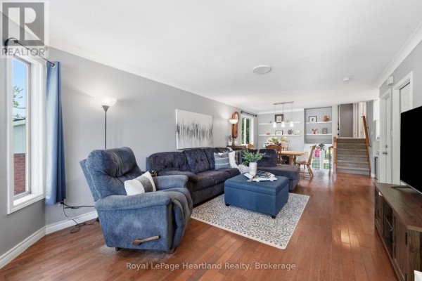 Living room with a sectional sofa, ottoman, and hardwood floors. Large window overlooks a treed area outside. - 54 Coleman Street, Huron East