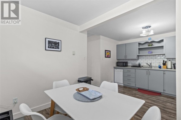 A dining area with a white table and four white chairs, adjacent to a kitchen with gray cabinets. Dark wood-look laminate flooring is visible. - 54 Nash Crescent, Mount Pearl