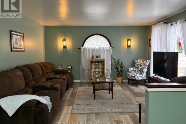 Living room with sage green walls, a dark brown sectional sofa, patterned armchair, and a television on a media stand. - 54 Redwing Ave, Manitouwadge