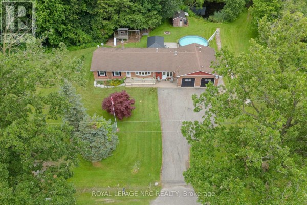 Aerial view of a residential property with a bungalow, garage, pool, and surrounding trees. - 54038 Willford Road, Wainfleet
