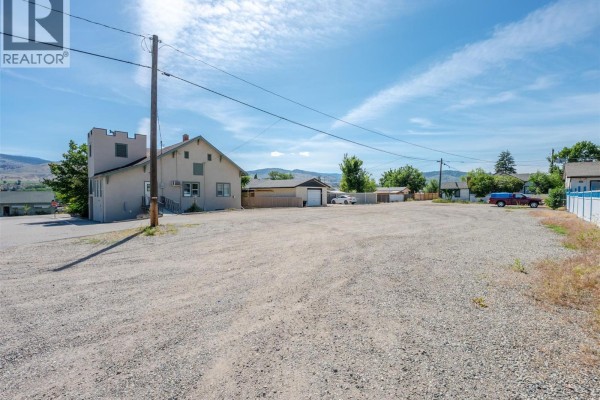 Gravel lot with a light-colored building featuring a castle-like tower, adjacent garages, and distant mountains. - 545 Church Avenue, Oliver