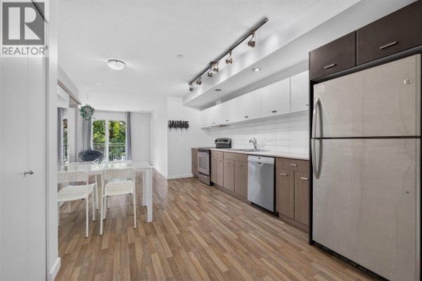 Open-concept kitchen and dining area with white counters, stainless steel appliances, and light wood flooring. - 547 Yates Road Unit# 211, Kelowna