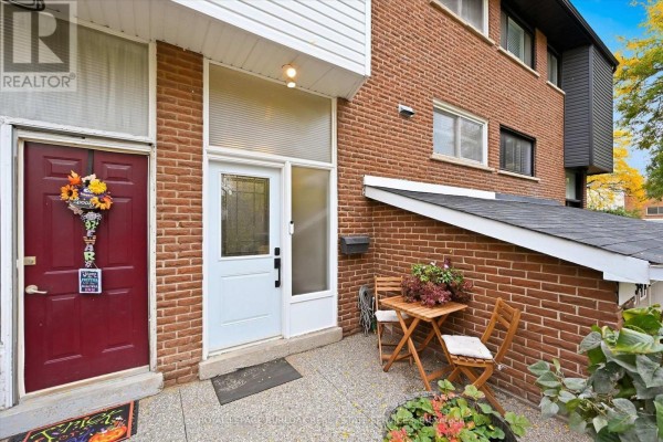 Front entrance of a brick townhouse with a red door, white door, and small patio table. - 5474 Schueller Crescent, Burlington
