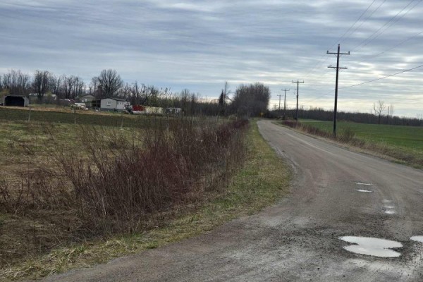 Dirt road curves past fields and houses with hydro poles under a cloudy sky. - 5496 Fairvalley Road, Ramara Road, Ramara