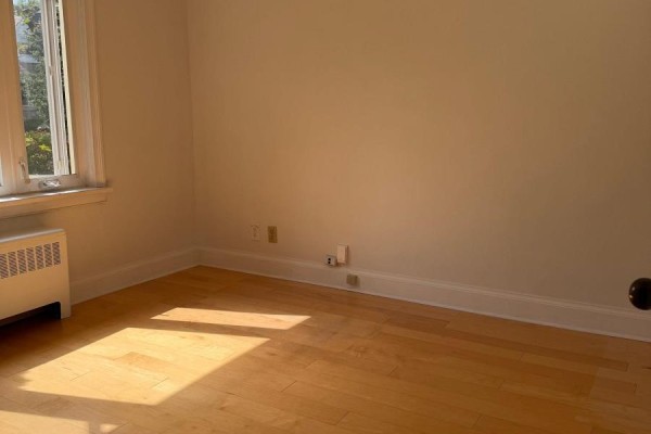 Empty room with hardwood floors, light beige walls, electrical outlets, and a radiator under a large window. - 55 Craigmore Crescent, Toronto